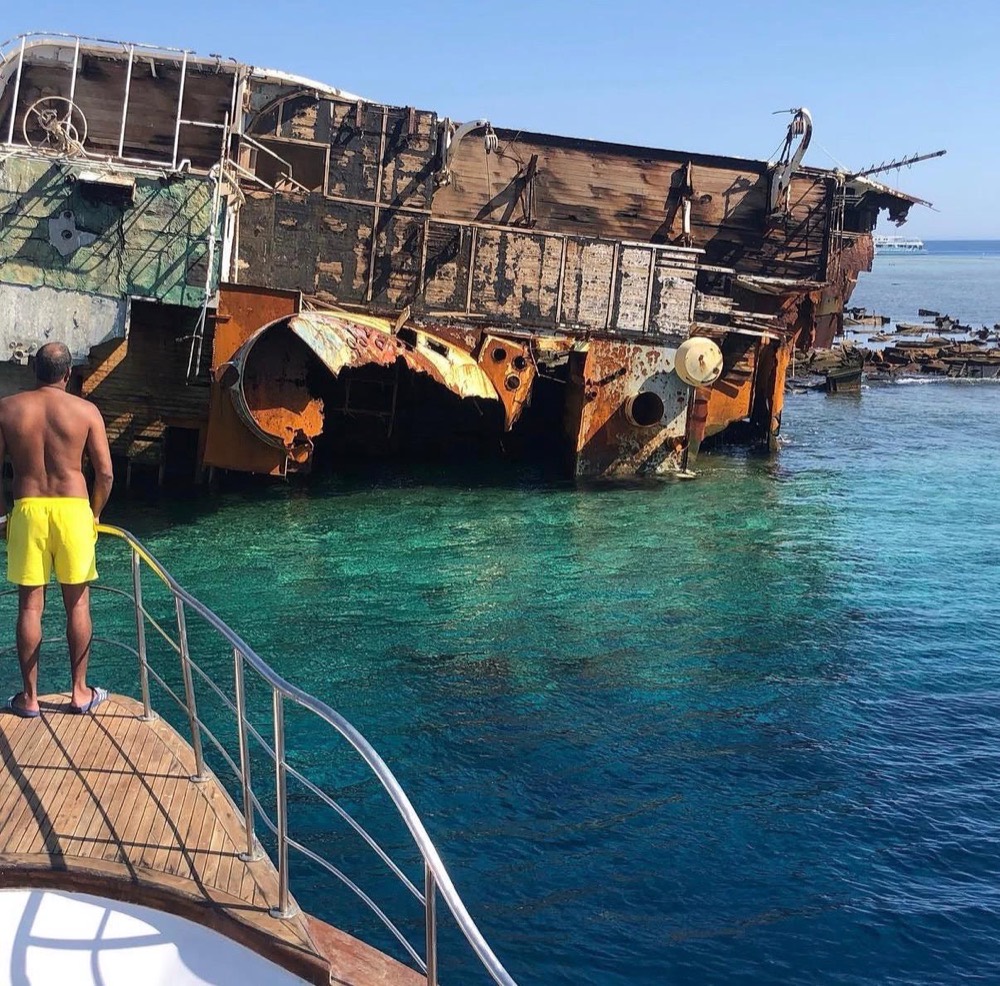 A guest stands on the bow looking at a large rusted shipwreck lying on a reef in clear blue-green water.