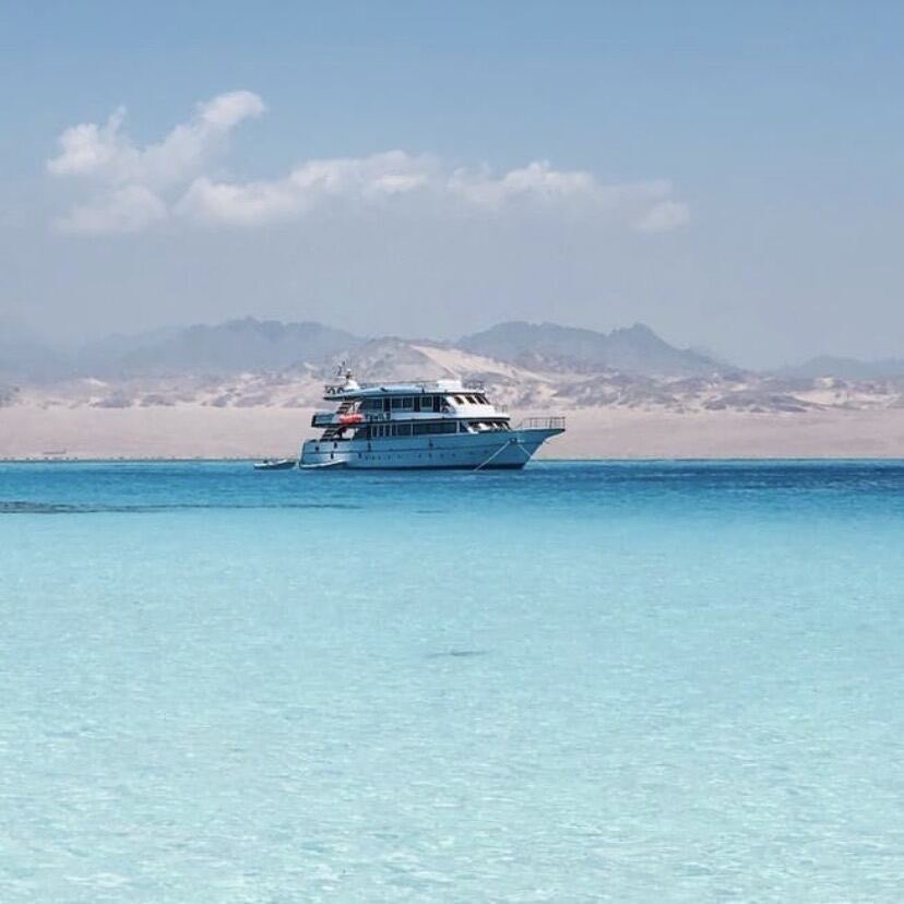 El Tawil 2, a white three-deck motor yacht, at anchor in turquoise shallows with desert mountains behind.